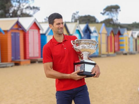 Novak Djokovic berdiri di depan beberapa gubuk pantai berwarna cerah dengan kemeja polo merah, memegang trofi Australia Terbuka