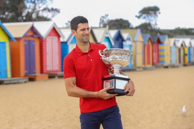 Novak Djokovic berdiri di depan beberapa gubuk pantai berwarna cerah dengan kemeja polo merah, memegang trofi Australia Terbuka
