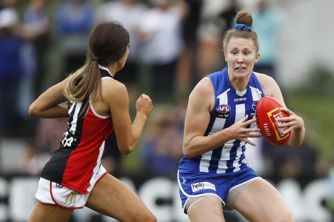 A Kangaroos AFLW player braces as she holds the football while a St Kilda player closes in on her.