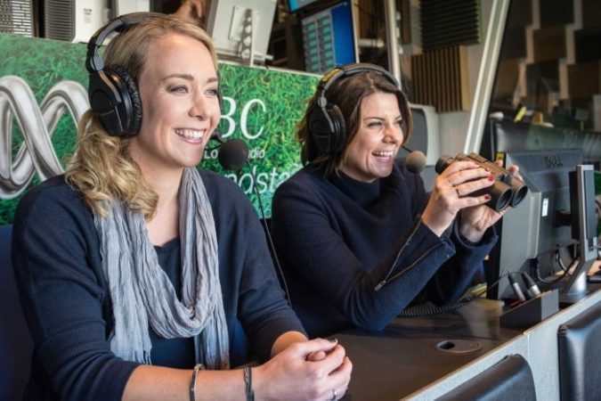 Two female broadcasters wearing headphones call an AFL match from a commentary box.