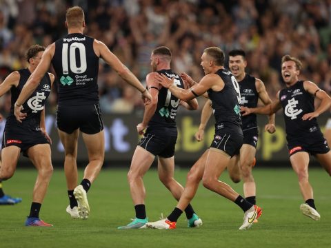 A group of AFL teammates grin widely as they rush in from all angles to get to their teammate who has just kicked a goal.