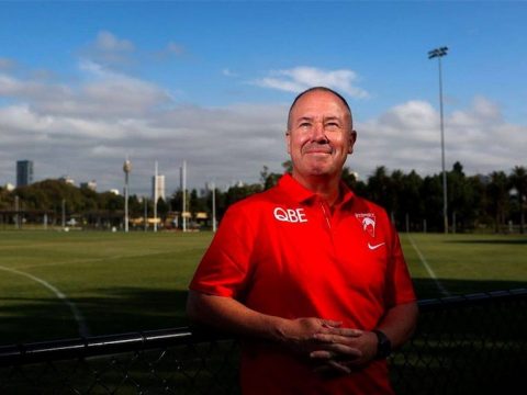 A man in a Sydney Swans polo smiles and looks at the sky in front of Sydney city