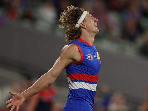 An AFL forward wearing a headband throws his head back and his arms wide after scoring a goal.