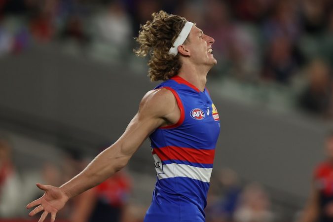 An AFL forward wearing a headband throws his head back and his arms wide after scoring a goal.