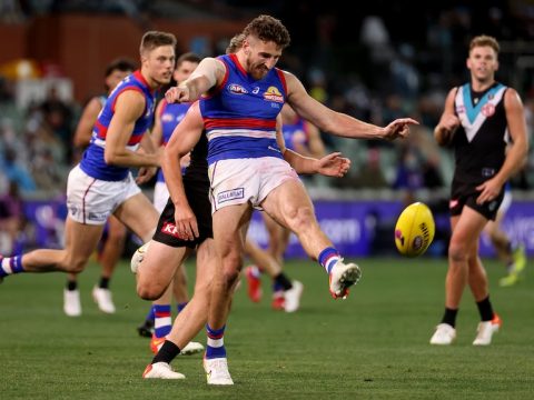 An AFL footballer extends his left leg in the act of kicking the football, as a defender trails him and another looks on.