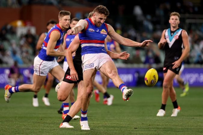 An AFL footballer extends his left leg in the act of kicking the football, as a defender trails him and another looks on.