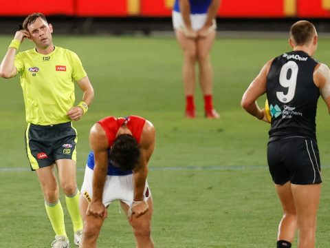 An AFL player bends over with his hands on his hips as an umpire signals behind him during a game.