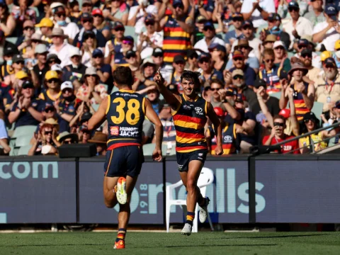 A young Crows AFL player points in the air in celebration after kicking a goal as a teammate runs towards him.