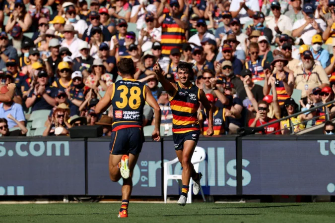 A young Crows AFL player points in the air in celebration after kicking a goal as a teammate runs towards him.