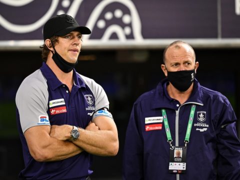 A star AFL footballer stands next to a coach with a frustrated expression and his arms folded  during a game.