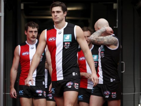 An AFL captain in red, black and white stands in the player's race before a game with his team behind him.