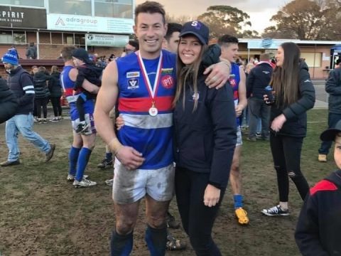 A man in muddy footy gear smiles while hugging his female partner, dressed in club merchandise.