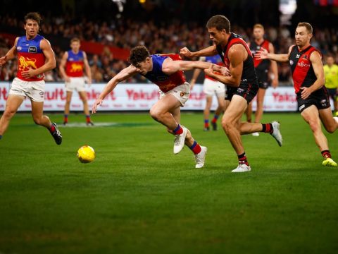 An AFL player puts his head down and reaches for the ball ahead of him, while a defender tries to hold his arm back.