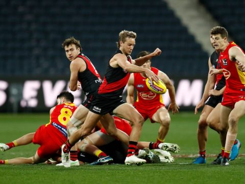 An Essendon midfielder looks at the ball as he kicks it downfield, while a group of other players lie on the ground behind him.