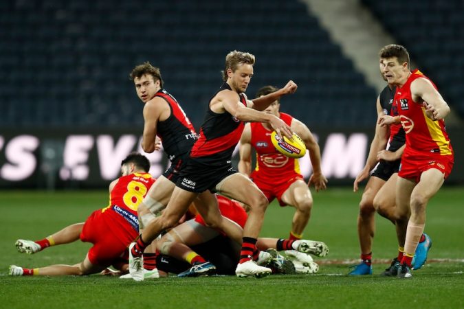 An Essendon midfielder looks at the ball as he kicks it downfield, while a group of other players lie on the ground behind him.