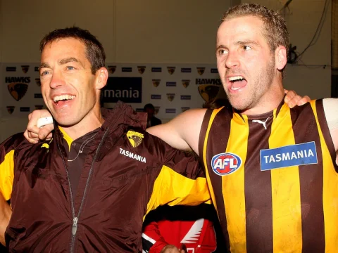 Hawthorn coach Alistair Clarkson stands with a beaming smile, singing, with his arm around the shoulder of one of his players.