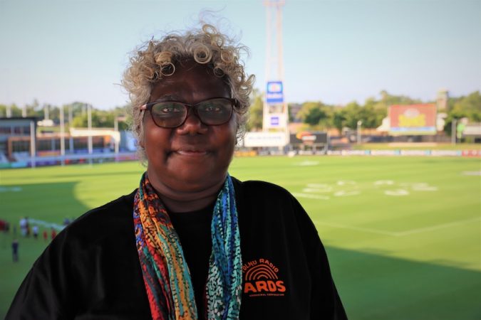 A woman with white curly hair smiles at the camera. Behind her is a football oval.