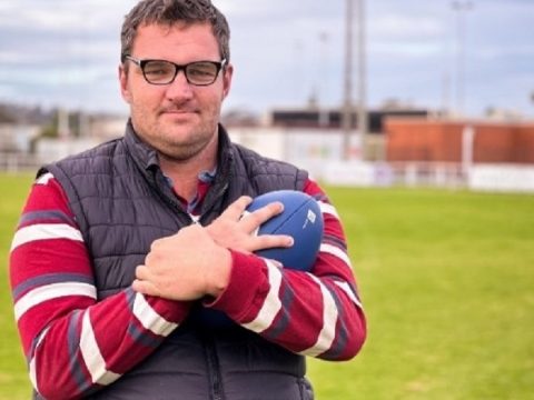 A man wearing glasses standing in a football field holding a football to his chest