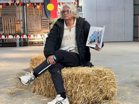 A woman with white hair and glasses and tracksuit sits cross legged on a hay bale holding a book.
