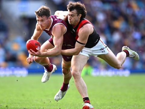 A Brisbane Lions AFL player attempts to handball while being tackled by an Essendon opponent.
