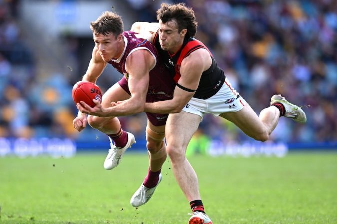 A Brisbane Lions AFL player attempts to handball while being tackled by an Essendon opponent.
