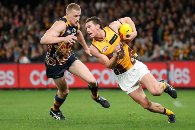 A Hawthorn AFL player holds the ball while being chased by an Adelaide opponent.