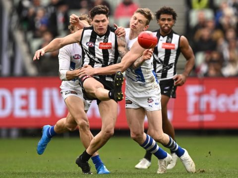 A Collingwood AFL player kicks the ball while being tackled by a North Melbourne opponent.