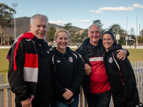 Two men and two women gather in front of a football oval and smile for a photo.