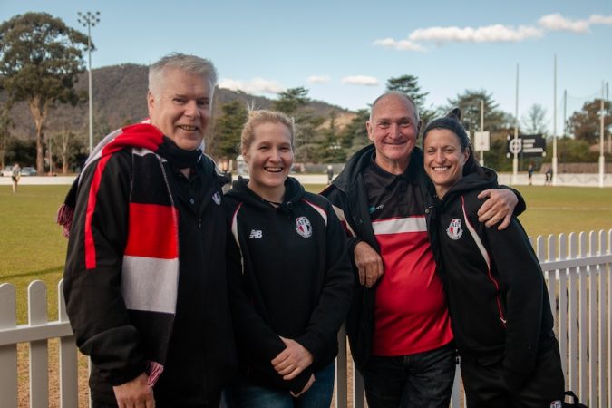 Two men and two women gather in front of a football oval and smile for a photo.