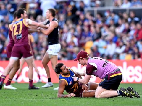 Callum Ah Chee is attended to by a medic while lying on the ground. Behind him, two Lions players wrestle Patrick Cripps