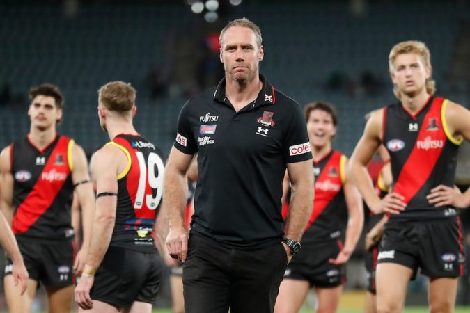 Essendon coach Ben Rutten looks stony-faced as his dejected players stand behind him after a big loss.