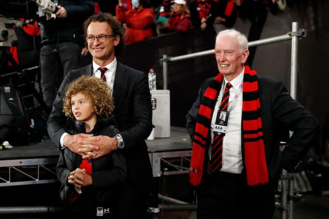 A man in a suit stands with his arms around a young girl, while next to him another man in a suit wearing an Essendon scarf.