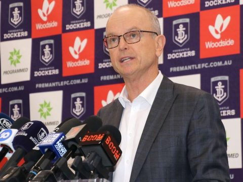 A mid-shot of Dale Alcock speaking in front of a board of Fremantle Dockers sponsor logos.