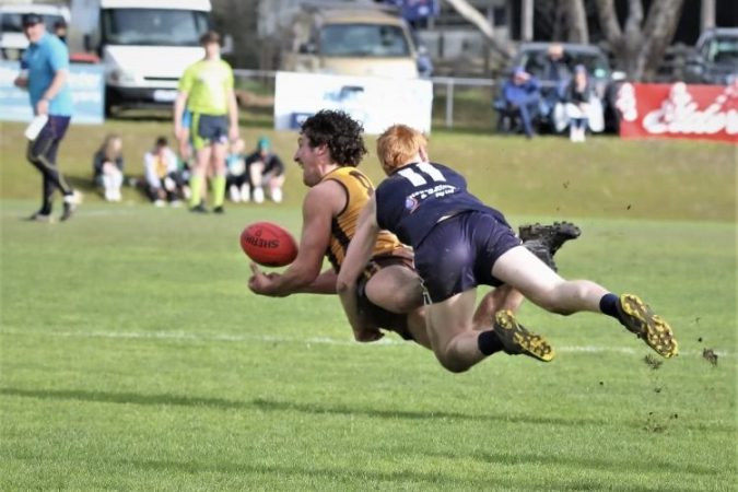 A football player in blue tackles another player in yellow on a footy ground as people watch on, cars in the distance.