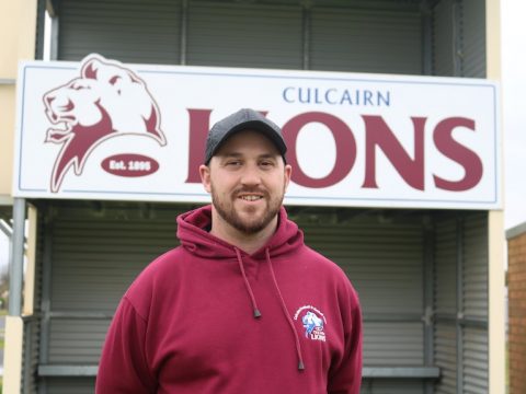 A man in a maroon jumper standing in front of the Culcairns Lions sign