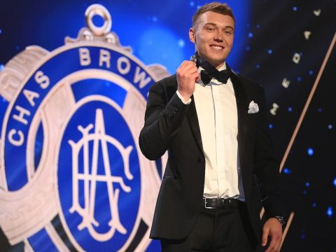 A Carlton AFL player stands on stage with a medal around his neck, and a sign behind him with the image of the Brownlow Medal.