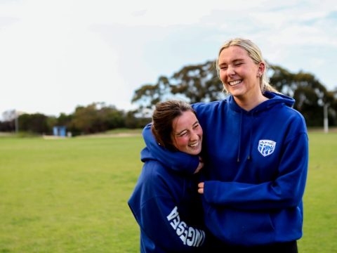 Two young women smile wrestling wearing blue hoodies on a football oval