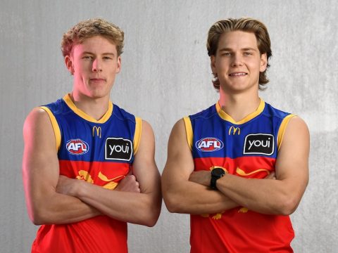 Two young men in Brisbane Lions guernseys stand facing the camera with arms folded.