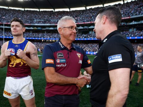 Two AFL coaches shake hands on the MCG after the grand final, while a losing player standing on the left of screen looks away.