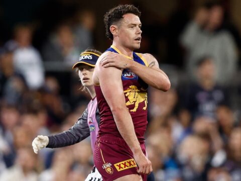 Brisbane Lions player Lachie Neale stands on the ground holding his sore shoulder as a team trainer watches.
