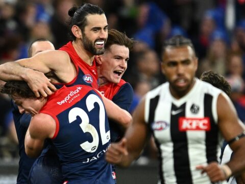 Three AFL players wearing red and blue, celebrate a goal, while an opposition play looks downcast