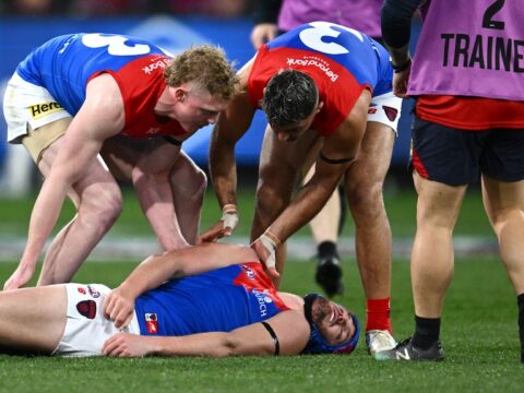 One AFL player lies motionless on the ground, two players at his aid, as a trainer runs on the ground.