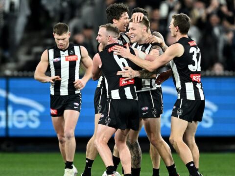 A group of Collingwood AFL players embrace and celebrate after a goal.
