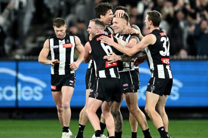 A group of Collingwood AFL players embrace and celebrate after a goal.
