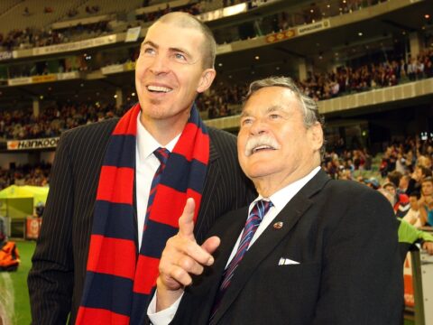 A tall man smiles wearing a Melbourne scarf around his neck as he stands next to an older man in a suit.