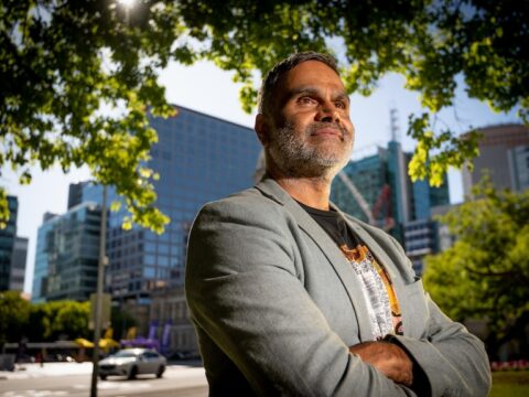A young Aboriginal man in a grey suit stands under a tree in front of some corporate buildings.