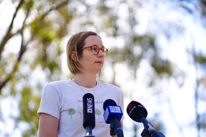 A woman wearing a tshirt with trees on it stands in front of media microphones with trees behind her, with a serious expression