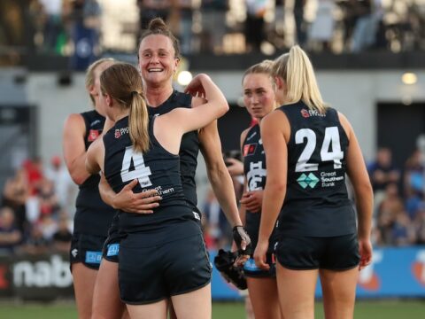 A group of Carlton AFLW players clad in navy blue grin and hug on the ground after a win.