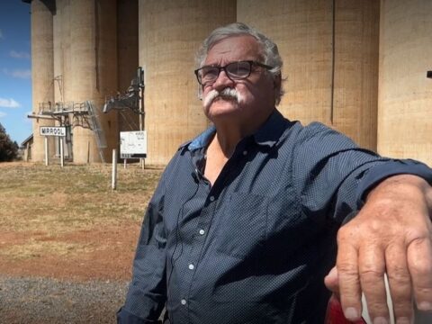A man with a moustache and black glasses sits in front of grain silos.